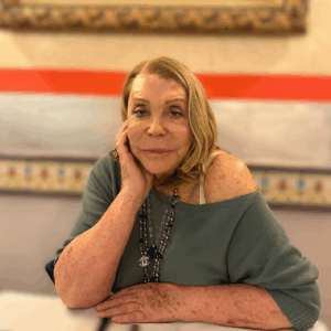 Portrait of Romina Cecconi with her chin resting on her hand, wearing a long necklace and a green boat neck jumper, indoors with a blurred background.
