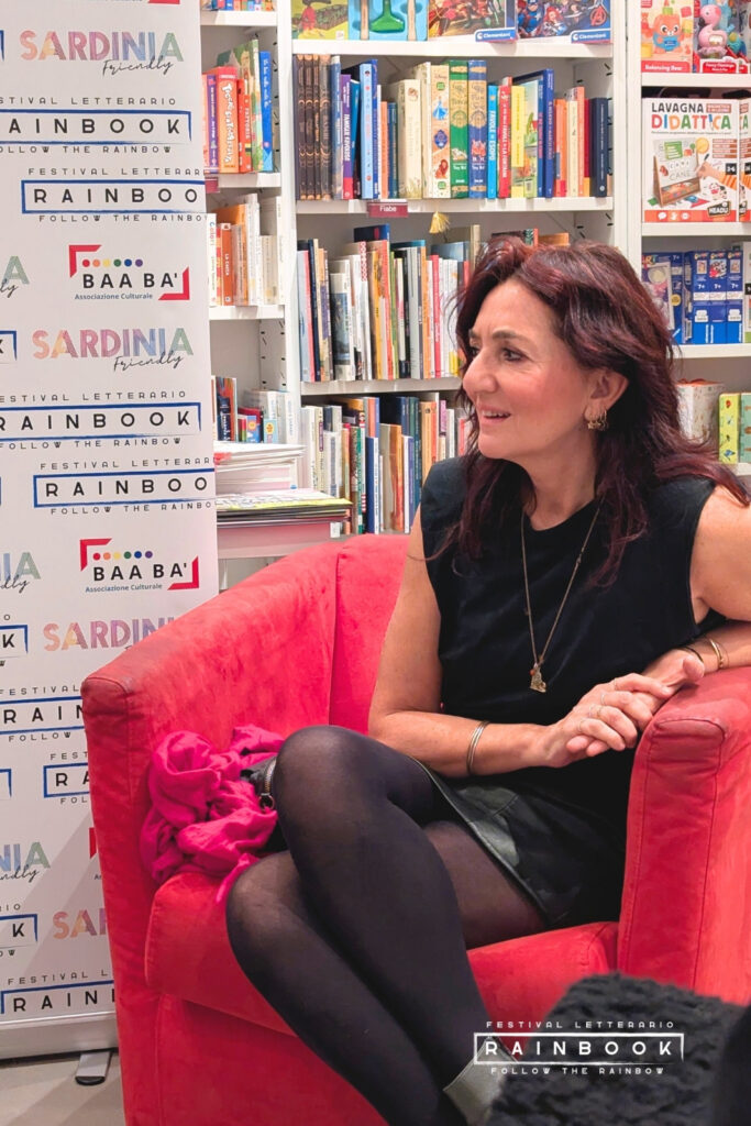 Vanessa Tonnini (Autrice) seduta in poltrona rossa durante il Festival Rainbook 2025, profilo laterale, mani intrecciate, collana lunga; sullo sfondo scaffali pieni di libri e banner con loghi Rainbook, BAABAI e Sardinia Friendly